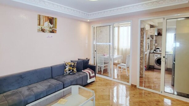 Ein Wohnzimmer mit grauem Sofa, einem Glastisch und einer Chandelier. Eine Glastür verschiebt sich zu einem Küchenbereich.