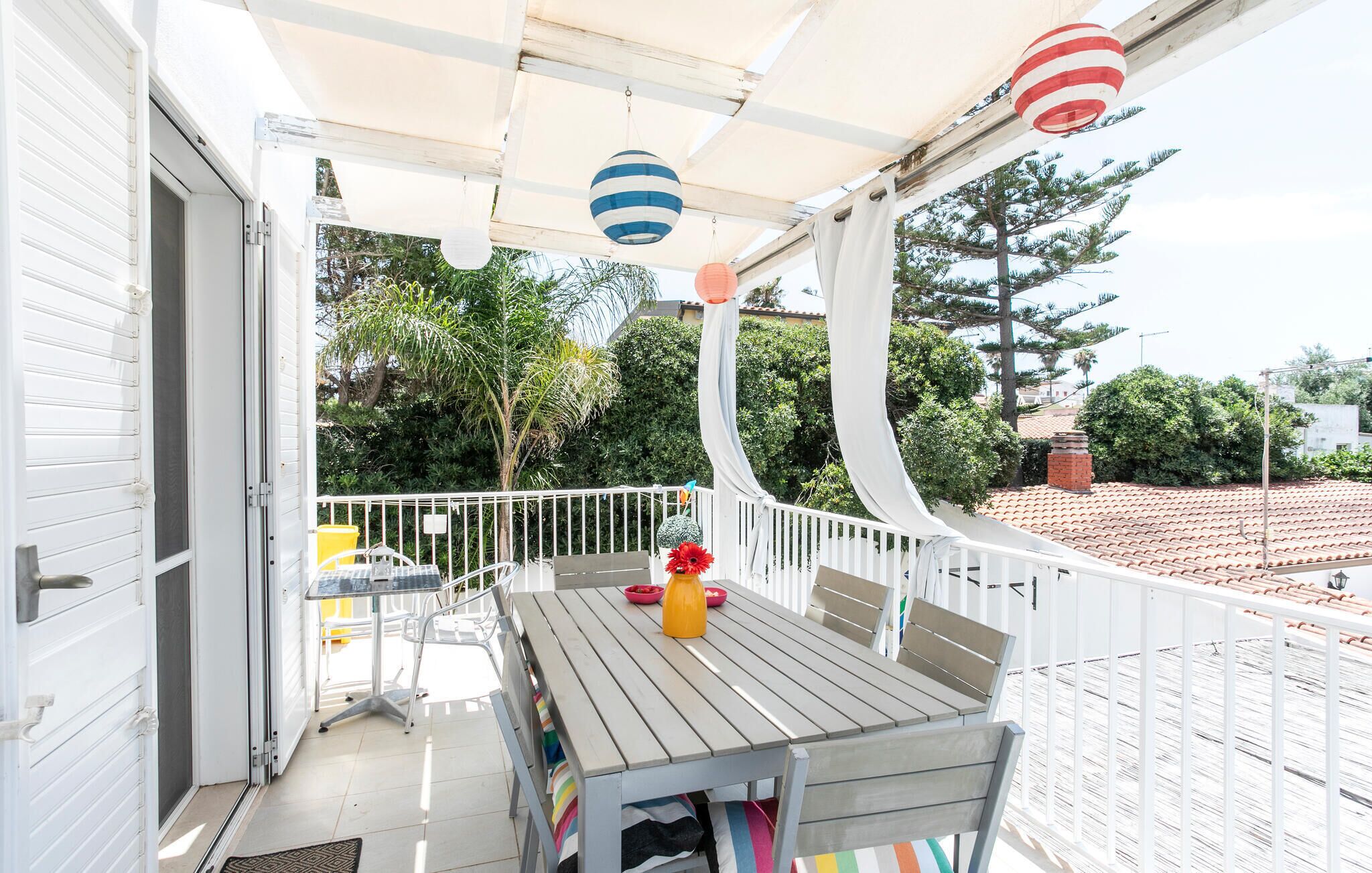 A covered outdoor patio with a wooden table and chairs, a yellow bucket, and a red flower vase.