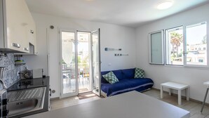 Et kompakt køkken med vask, skabe og et spiseområde. Der er en blå sofa og et lille bord i opholdsstuen.