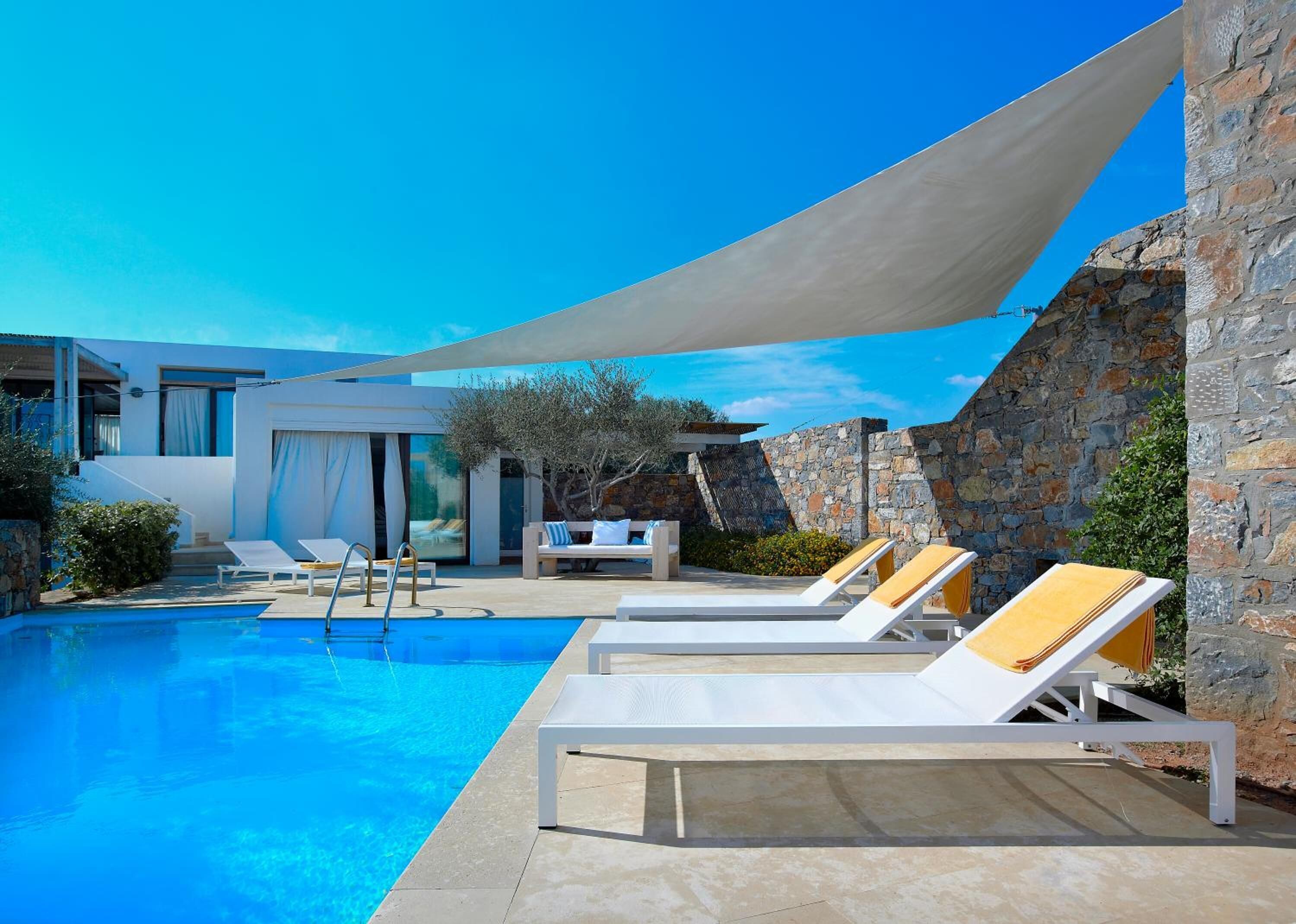 Uma área de piscina moderna, com espreguiçadeiras brancas e um muro de pedra ao fundo.