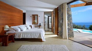 A bedroom with a large bed, a nightstand, a mirror, and a view of a pool and ocean. - Thalassa Villas (Agios Nikolaos)