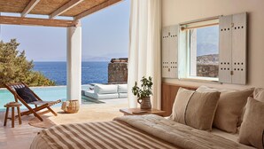 A bedroom with a large bed, a view of the sea, a wooden chair, and a small table with a plant. - Thalassa Villas (Agios Nikolaos)