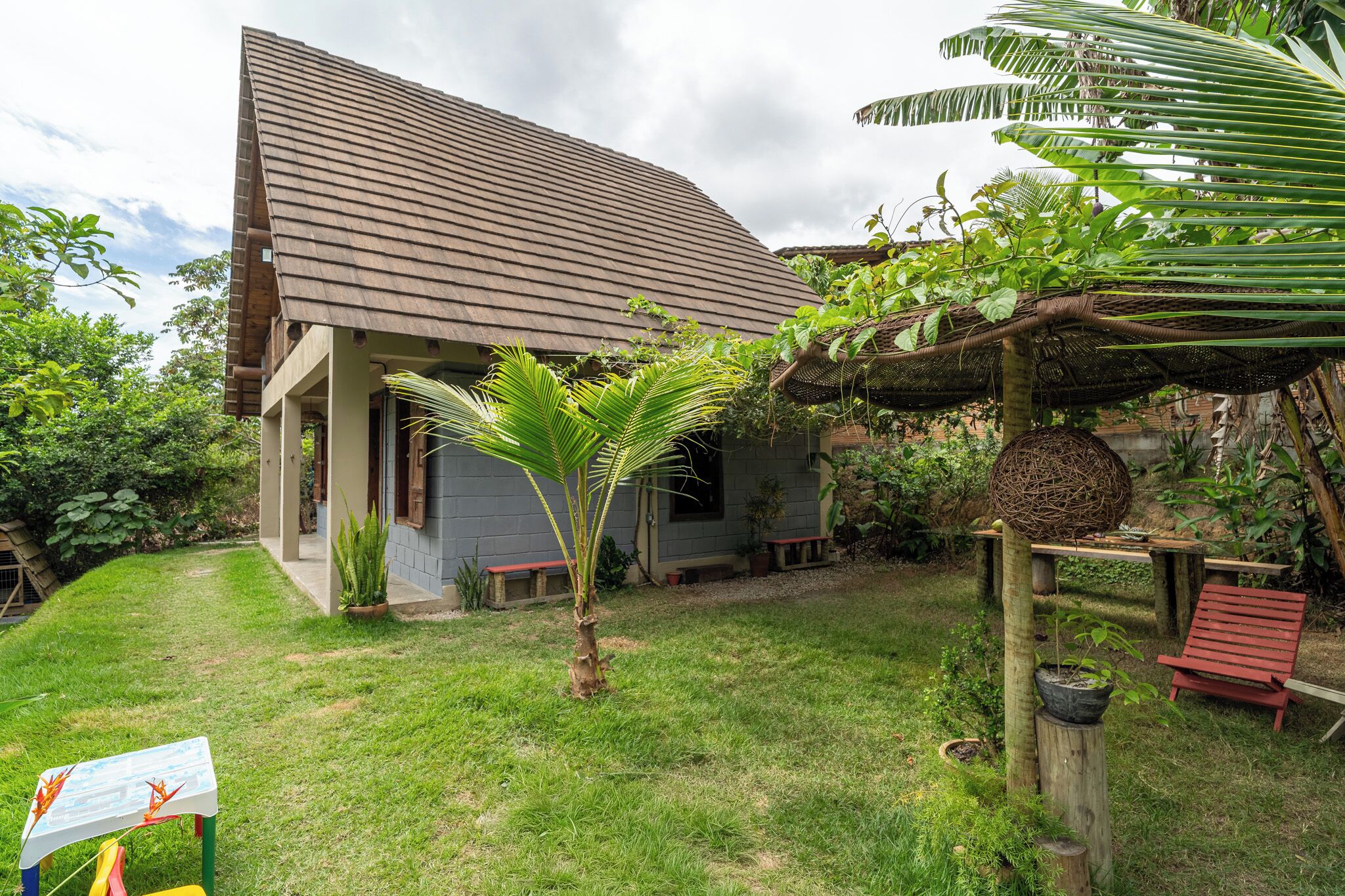 Een huis met een schuin dak, een klein terras en een tuin met een palmboom en een houten schommel.