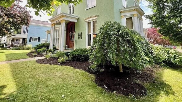 A two-story house with a front porch, a well-maintained lawn, and a large bush in the front yard. - Wisteria Suite -The Belvedere Inn (Oswego)