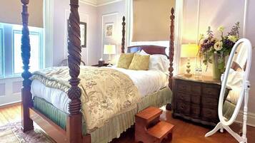 A four-poster bed with a floral bedspread, a dresser with a mirror, and a small wooden stool.