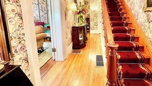 A grand hallway with a red carpeted staircase, floral wallpaper, and a wooden floor. - Wisteria Suite -The Belvedere Inn (Oswego)