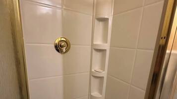 Un box de ducha pequeño y cerrado con azulejos blancos, un grifo de ducha dorado y un estante sencillo.