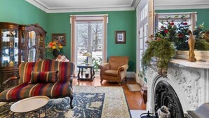 A cozy living room with a striped sofa, a wooden cabinet, a fireplace, and a large window. - Gardenia Suite - The Belvedere Inn (Oswego)