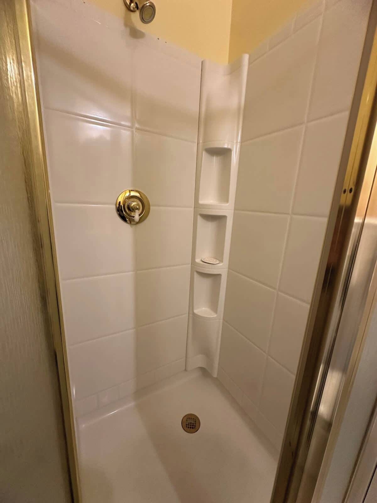 A small, enclosed shower stall with white tiles, a gold shower knob, and a single shelf.