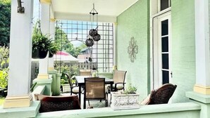 A covered patio with a table and chairs, a decorative wall, and a window with a metal grid. - Gardenia Suite - The Belvedere Inn (Oswego)
