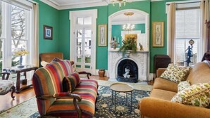 A room with a fireplace, a striped armchair, a sofa, and a chandelier. - Gardenia Suite - The Belvedere Inn (Oswego)