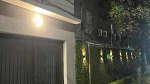 Un edificio con entrada protegida por una reja, un muro cubierto de vegetación y un sendero que conduce a una zona vallada.