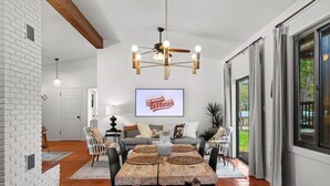 A modern living room with a dining area, featuring a fireplace, a large TV, and a ceiling fan. - * Stunning FBG Retreat * | Sleeps 10 (Fredericksburg)