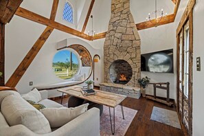 Un salon chaleureux avec une cheminée en pierre, un grand téléviseur et une fenêtre circulaire donnant sur un paysage magnifique.