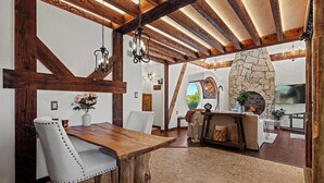 Un intérieur rustique avec une cheminée en pierre, des poutres en bois et un coin repas comprenant une grande table en bois et des chaises blanches.