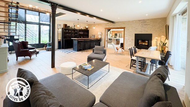 Un salon moderne avec un canapé, une table basse et un coin repas. Au fond, on aperçoit une cuisine avec un bar et une cheminée.