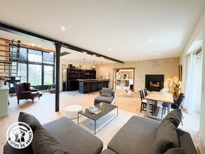 Ein modernes Wohnzimmer mit Sofa, Couchtisch und Essbereich. Im Hintergrund befindet sich eine Küche mit Bar und Kamin.