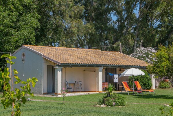 A single-story house with a tiled roof, a covered patio with a table and chairs, and a lawn with two lounge chairs. - Lefkes Apartments (Corfu)