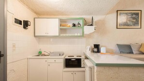 A compact kitchen with white cabinets, a microwave, and a small countertop. There is a shelf above the sink with a kettle and cups. A painting hangs on the wall, and a sofa with cushions is visible in the background. - Lefkes Apartments (Corfu)