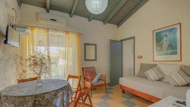A cozy living room with a sofa, a dining table with chairs, a small table with papers and a remote, and a door. - Lefkes Apartments (Corfu)