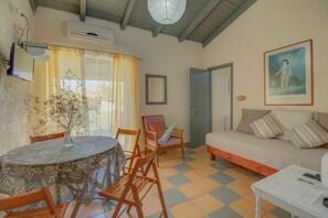 A cozy living room with a sofa, a dining table with chairs, a small table with papers and a remote, and a door. - Lefkes Apartments (Corfu)