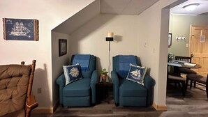 En stue med to blå lænestole, en brun lænestol, et billedrammet skibsbillede, en lampe og et spiseområde i baggrunden.