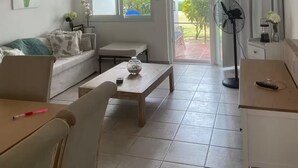 A living room with a sofa, coffee table, and a fan. There is a window with a view of greenery outside. - Beach Apartment in Stanza Mare Punta Cana (Punta Cana)
