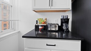A kitchen with white cabinets, a black countertop, a toaster, a coffee maker, and a window with blinds. - Stunning City Views · Midtown Heart of Detroit · Clean · Free Parking (Detroit)