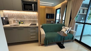 A modern kitchen with a microwave, sink, and storage cabinets. There is a green sofa with a blanket and pillows, and a window with curtains.