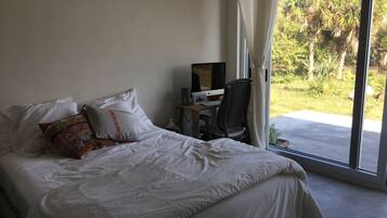 A bedroom with a bed, a desk with a computer, and a view of greenery outside.