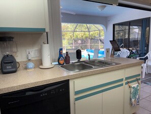 Een keuken met een vaatwasser, een blender, een keukenrolhouder, een fles afwasmiddel en een raam met uitzicht op een zwembad en groen.