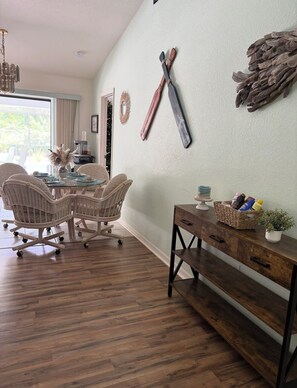 Een eetgedeelte met een glazen tafel, rieten stoelen, een houten bijzettafeltje met een mand en een rustiek decoratief object aan de muur.