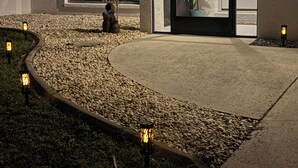 Entrada de un edificio con un camino curvo, plantas en macetas e iluminación exterior.