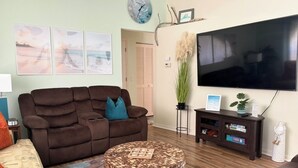 Una sala de estar con un sofá marrón, una mesa redonda de centro, un televisor de pantalla plana y un reloj de pared.