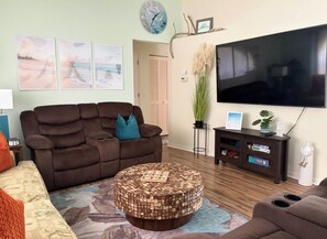 Een woonkamer met een bruine bank, een ronde salontafel, een flatscreen televisie en een wandklok.