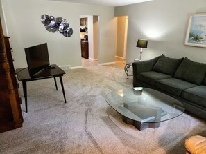 Ein Wohnzimmer mit Glastisch, dunklem Sofa, Fernseher auf einem Standfuß und einer dekorativen Metallskulptur an der Wand.
