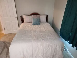 Ein ordentlich bezogenes Bett mit weißer Decke und gemusterten Kissen, ein Holz-Kopfstück und eine Tür links.