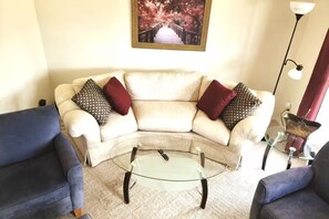 Una sala de estar con un sofá color beige, dos sillones, una mesa de centro de vidrio y una lámpara de pie.