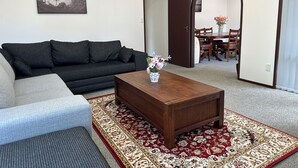 Una sala de estar con un sofá oscuro, un sillón de color claro, una mesa de centro de madera con un jarrón de flores y una alfombra estampada.