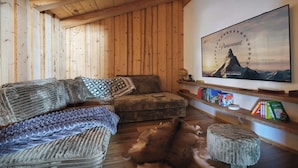 Een gezellige kamer met houten wanden, een grote flatscreen televisie aan de muur, een bank met gestreepte plaids, een leren vloerkleed en een boekenkast vol boeken en decoratieve objecten.