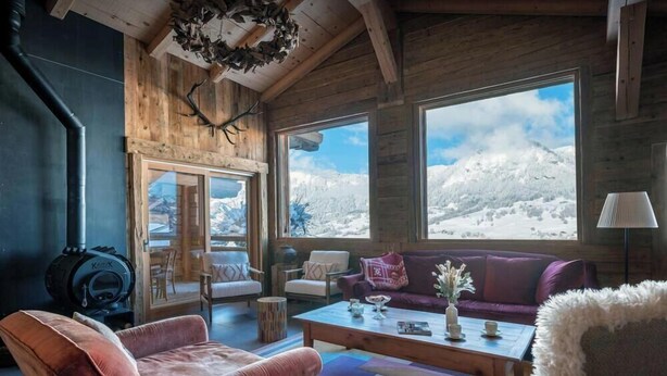 Een gezellig houten blokhutinterieur met een open haard, houten wanden, grote ramen en uitzicht op met sneeuw bedekte bergen.