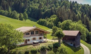 A two-story house with a large deck, surrounded by lush greenery and a forested hillside. - Ferme Claudius - Enjoy skiing and nature right on the piste - OVO Network (Praz-sur-Arly)