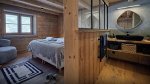 Een houten kamer met een bed, een raam, een badkamer met een wastafel en spiegel, en een kleine zithoek.