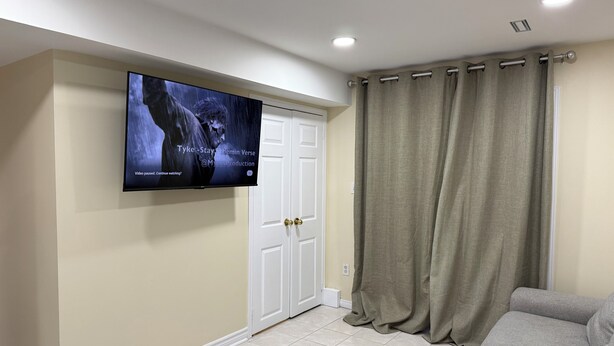 Una sala de estar con un televisor de pantalla plana montado en la pared, un sofá gris, paredes color beige, una puerta blanca y una ventana con cortinas beige.