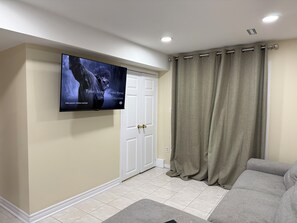 Un salon avec un téléviseur à écran plat fixé au mur, un canapé gris, des murs de couleur beige, une porte blanche et une fenêtre avec des rideaux beiges.