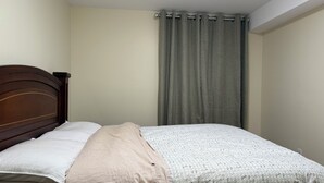 Un dormitorio con cabecera de madera, cama con colcha estampada, almohada color beige y cortina gris.