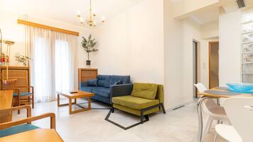Ein modernes Wohnzimmer mit einem blauen Sofa, einem grünen Sessel, Holzmöbeln und einem Essbereich mit weißen Stühlen.