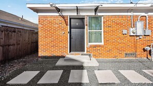 A brick building with a metal door, a small window with a green pattern, and a concrete step leading to a gravel area. - Cityscapes Studio Suite #D @ East Downtown (Houston)