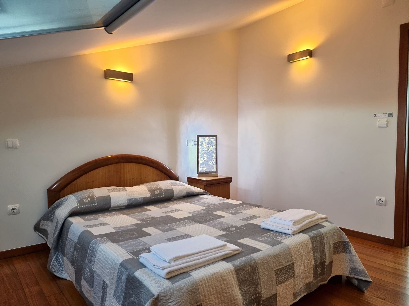 A neatly made bed with a quilt and folded towels, a wooden headboard, a bedside table with a lamp, and a window with a view of trees.