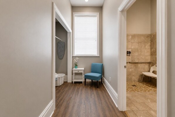 A narrow hallway with a blue chair, a small table with a plant, a basket, and a window with blinds.